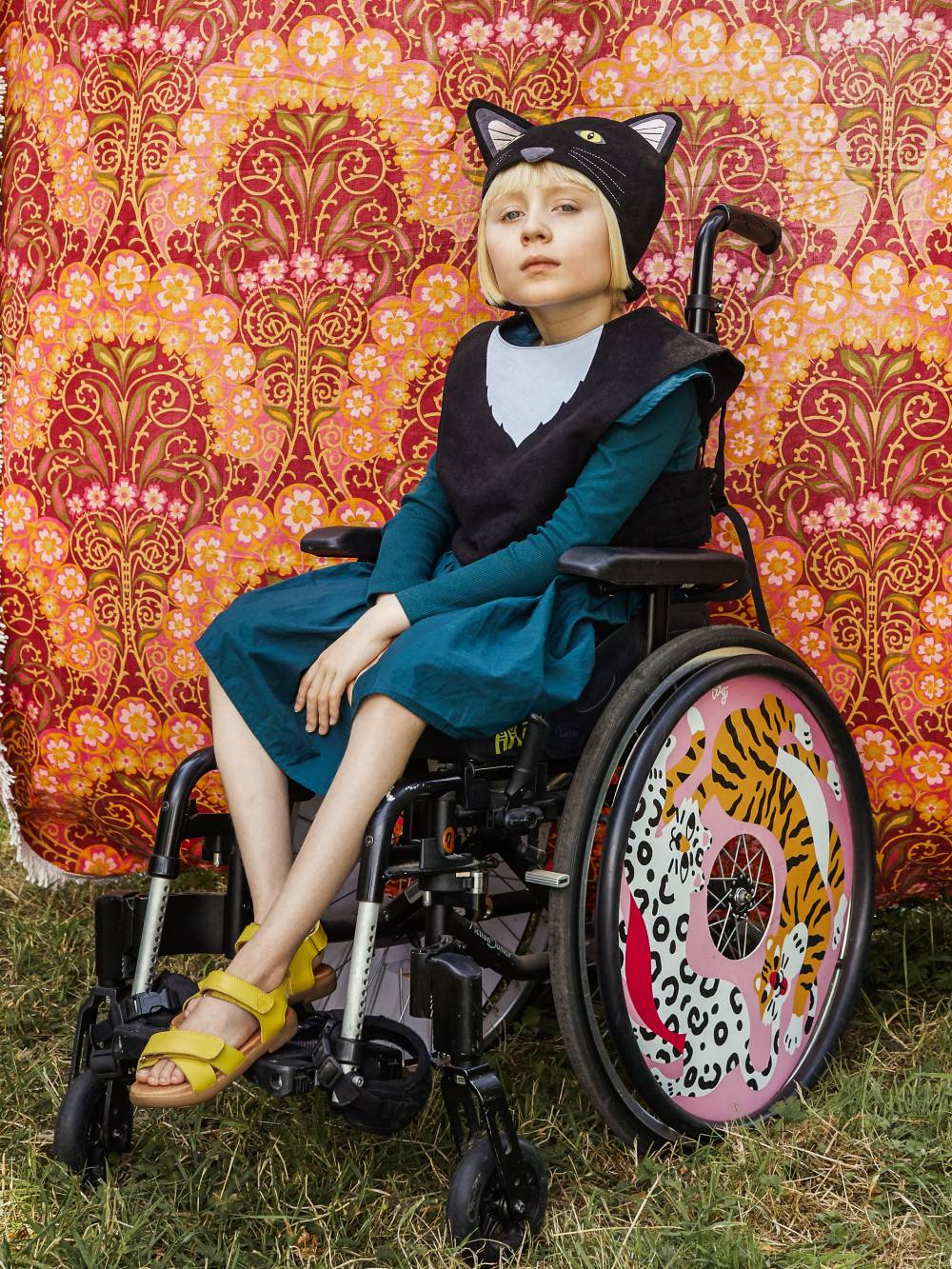 Girl wearing the black cat costume sitting in her wheelchair against a pink floral fabric backdrop.