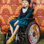 Girl wearing the black cat costume sitting in her wheelchair against a pink floral fabric backdrop.