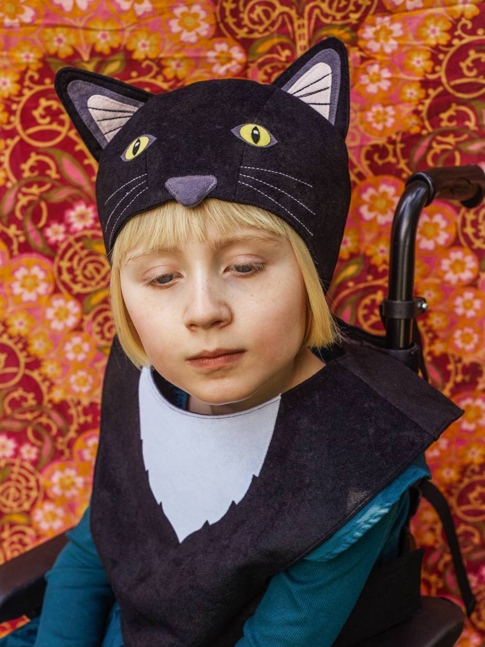 Girl wearing the black cat hat and tabard, sitting in her wheelchair against a pink floral fabric backdrop.