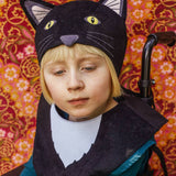 Girl wearing the black cat hat and tabard, sitting in her wheelchair against a pink floral fabric backdrop.