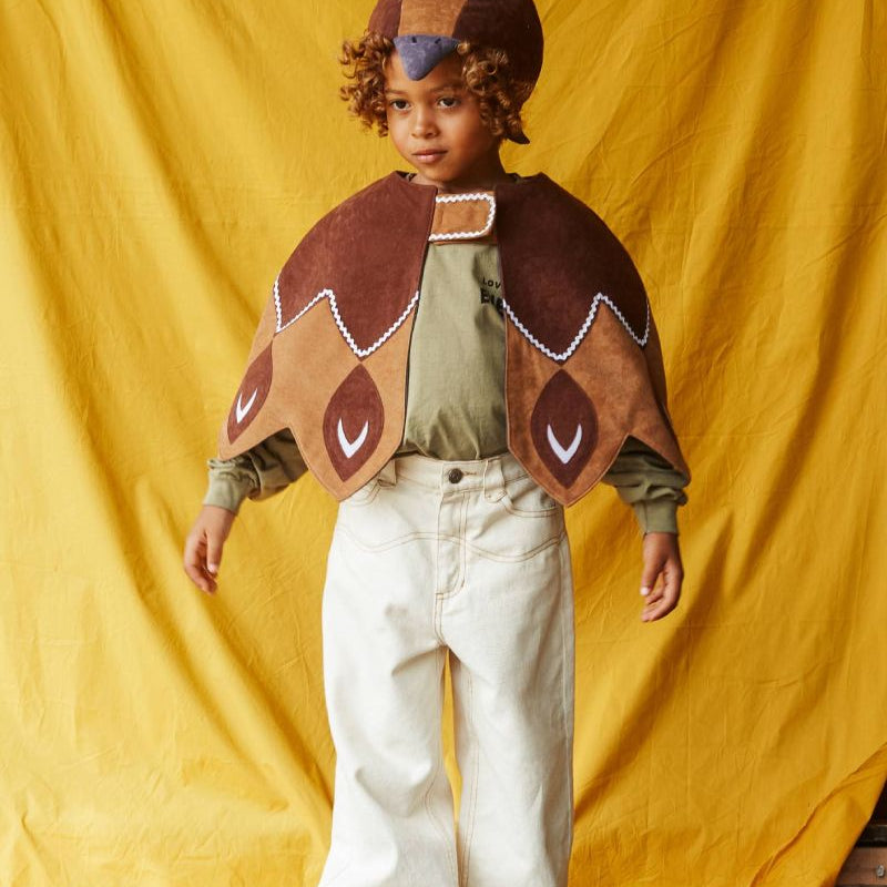 Child wearing the brown duck hat and brown feathered bird cape, standing in front of a yellow fabric background.
