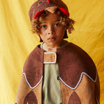 Child wearing the brown hen hat and brown feathered bird cape, standing against a bright yellow fabric background.