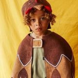 Child wearing the brown hen hat and brown feathered bird cape, standing against a bright yellow fabric background.