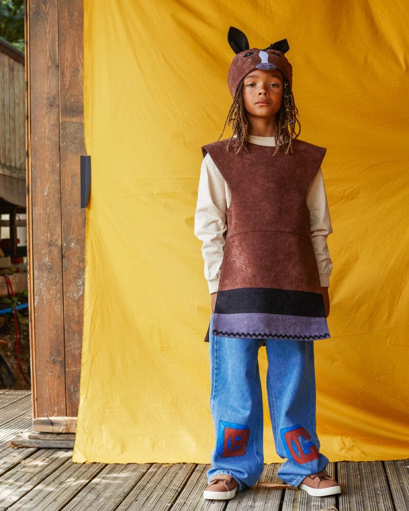 Child wearing the brown horse hat and tabard, standing in front of a bright yellow fabric background.