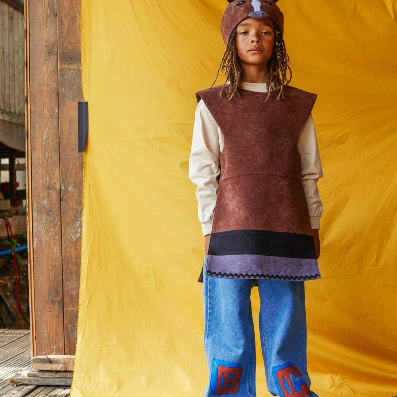 Child wearing the brown horse hat and tabard, standing in front of a bright yellow fabric background.