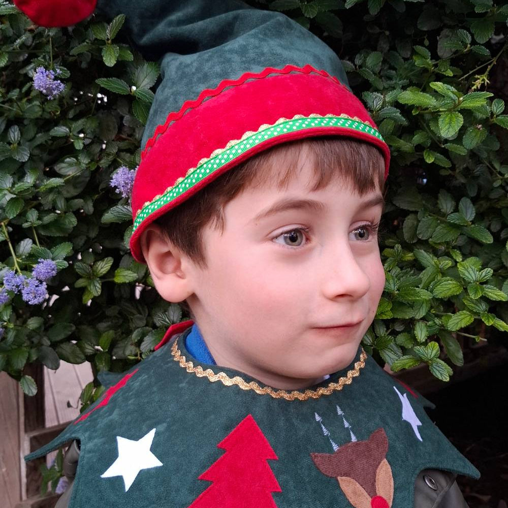 Head and shoulders of child wearing the woodland green Kids Christmas Elf Hat and woodland green Kids Christmas Costume Collar. He is smiling and standing against a green woodland background.