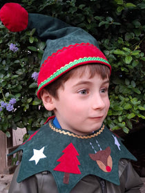 Head and shoulders of child wearing the woodland green Kids Christmas Elf Hat and woodland green Kids Christmas Costume Collar. He is smiling and standing against a green woodland background.