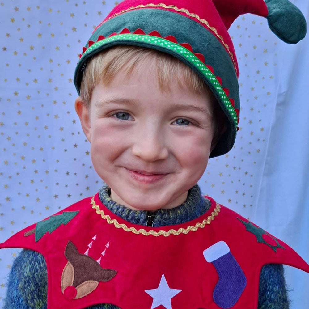 Head and shoulders of child wearing the red Kids Christmas Elf Hat and red Kids Christmas Costume Collar. He is smiling against a white and gold starry fabric backdrop.