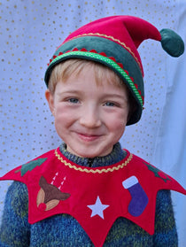Head and shoulders of child wearing the red Kids Christmas Elf Hat and red Kids Christmas Costume Collar. He is smiling against a white and gold starry fabric backdrop.
