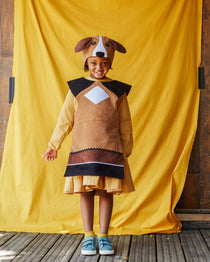 Child wearing the floppy eared dog hat and tabard, smiling at the camera. She is standing in front of a yellow fabric background.