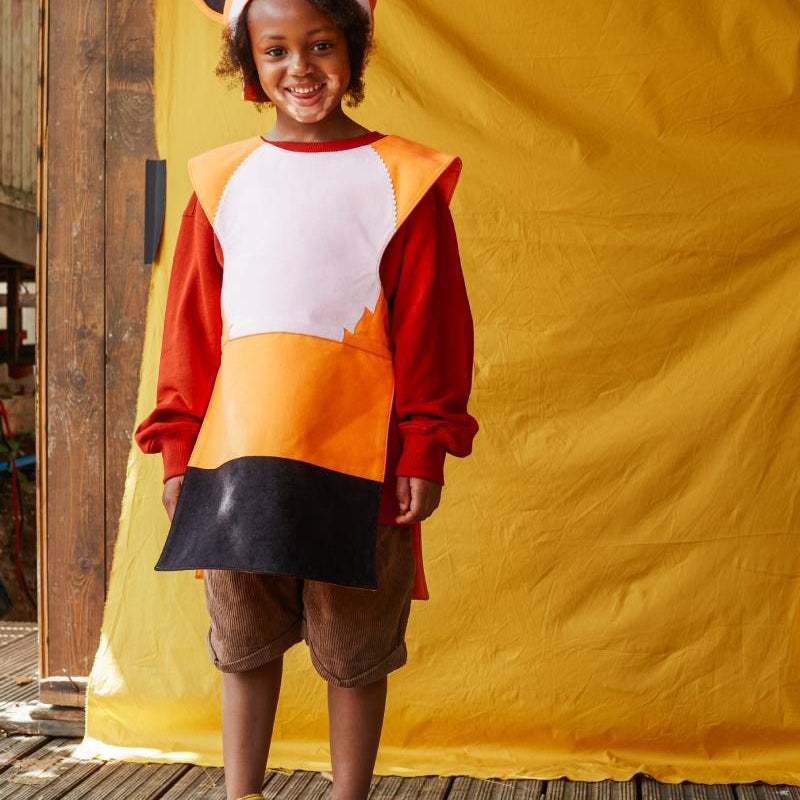 Child wearing the red fox costume smiling at the camera and standing against a yellow fabric background.