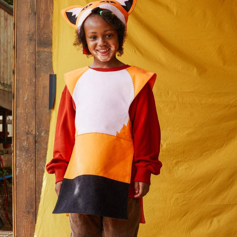 Child wearing the red fox hat and tabard smiling at the camera in front of a yellow fabric background.