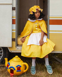 Girl smiling, sitting in the doorway of a caravan and wearing the white & yellow mob cap, reversible cap and yellow pinafore dress. There is a large bright yellow toy teapot by her feet.