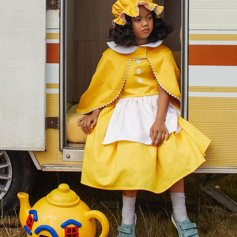 Girl smiling, sitting in the doorway of a caravan and wearing the white & yellow mob cap, reversible cap and yellow pinafore dress. There is a large bright yellow toy teapot by her feet.