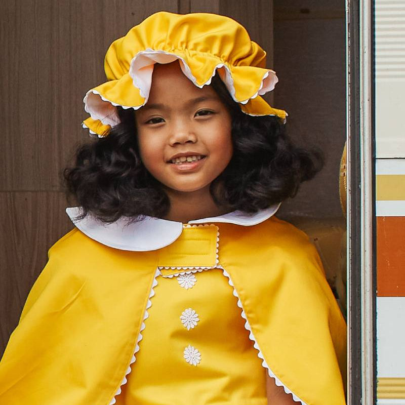 Girl smiling, sitting in the doorway of a caravan and wearing the white & yellow mob cap, reversible cap and yellow pinafore dress.