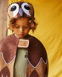 Child wearing the tawny owl hat and brown feathered bird cape. He is standing in front of a yellow fabric background.