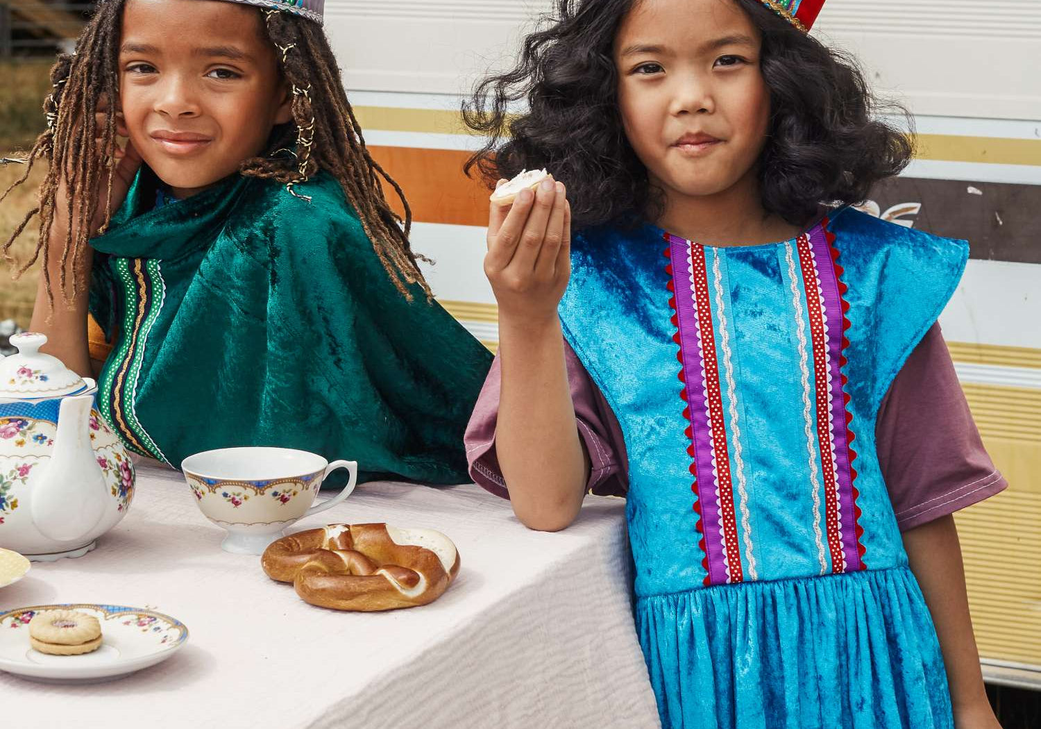 Girl wearing the turquoise royal fairytale dress and red fairytale crown eating a cake at a teaparty. To the left is a boy wearing the emerald fairytale cape and silver fairytale crown