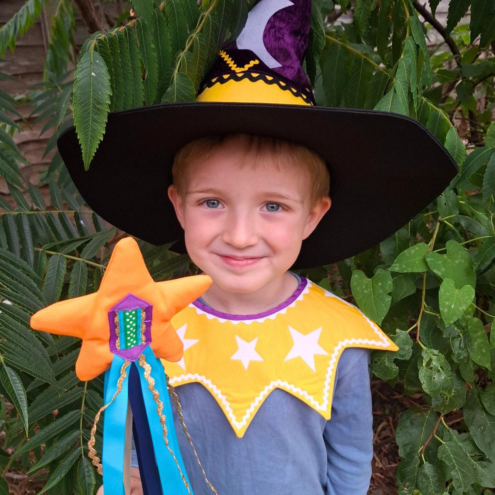 Smiling child wearing the twinkling stars kids costume collar, magical witch hat and holding the orange fairytale star wand, sitting outdoors.