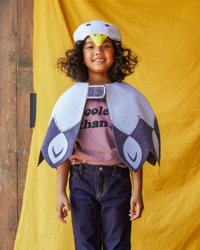 Child wearing the white feathered bird cape standing in front of a yellow fabric background.