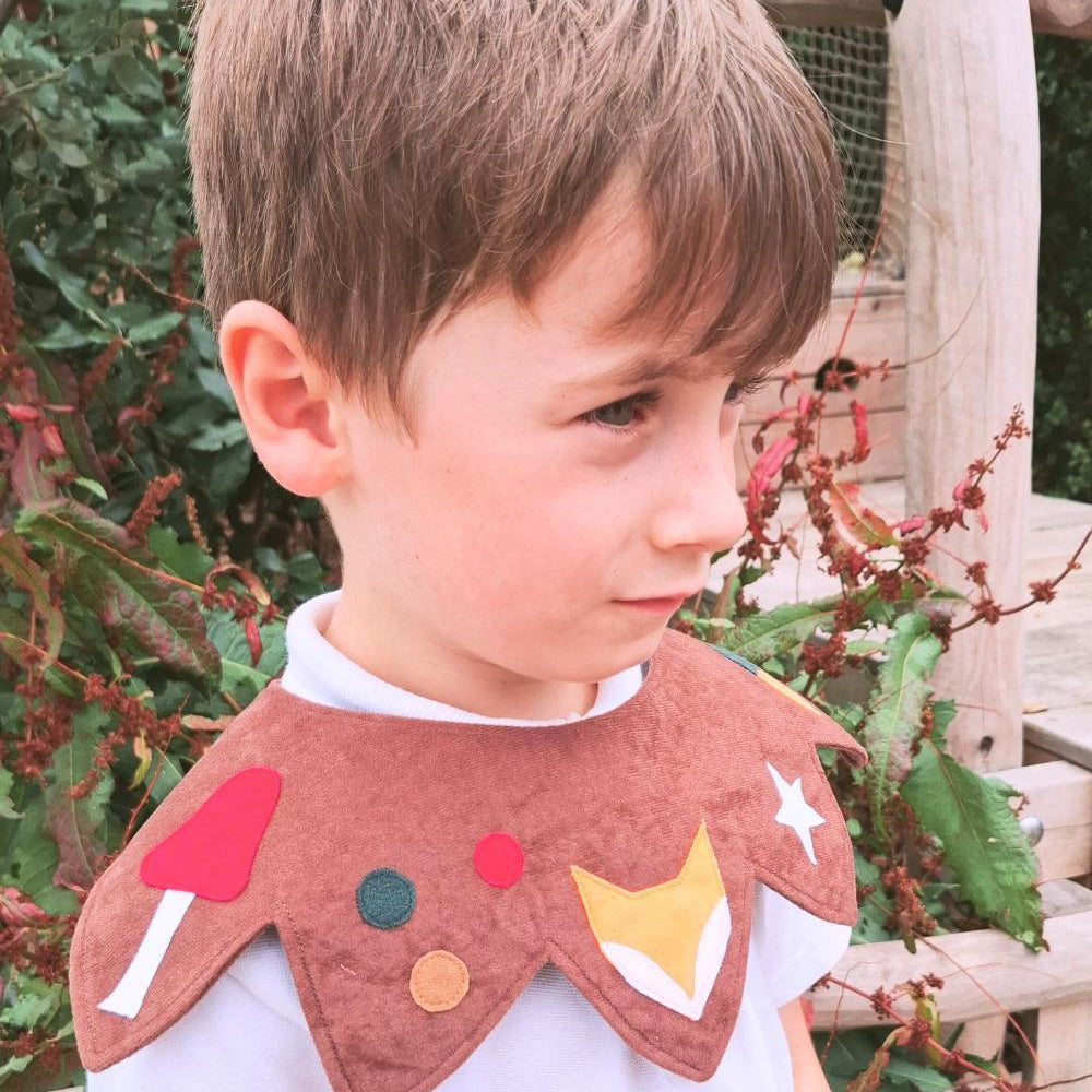 Boy standing in a garden, looking sideways, wearing the brown-coloured woodland kids costume collar over a white polo shirt.