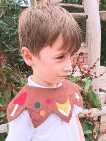 Boy standing in a garden, looking sideways, wearing the brown-coloured woodland kids costume collar over a white polo shirt.