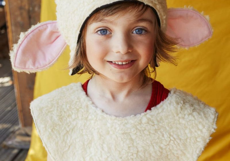 Child smiling at the camera wearing the woolly sheep hat and tabard, standing in front of a yellow fabric backdrop.